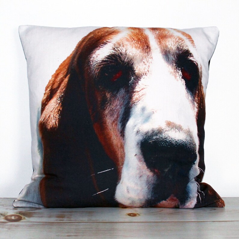 K&ouml;nnte beinhalten: Ein wei&szlig;es Kissen mit dem Aufdruck eines braunen und wei&szlig;en Basset-Hundegesichts. Der Hund hat einen ernsten Ausdruck und die Augen sind geschlossen.