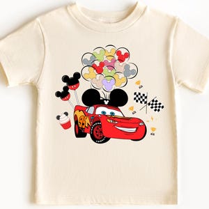 K&ouml;nnte beinhalten: Cremefarbenes kurz&auml;rmeliges T-Shirt mit einem roten Rennwagen mit der Nummer 95, einem l&auml;chelnden Gesicht und Mickey Mouse-Ohren. Das Design umfasst bunte Luftballons, karierte Flaggen und kleine Troph&auml;en.
