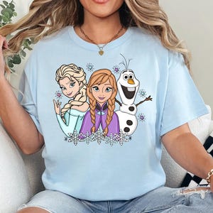 以下が含まれることがあります： アナと雪の女王のエルサ、アナ、オラフのグラフィックプリントが施されたライトブルーの半袖Tシャツ。キャラクターの周りには小さなカラフルな星が散りばめられ、胸元のイラストの下部には白い雪の結晶のデザインが描かれています。
