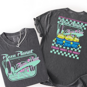 May include: Two dark gray t-shirts with Pizza Planet graphics. One shirt features a rocket and space diner design, the other has three green aliens and the text "Friday Night Kid Night". A silver chain necklace is on one shirt.