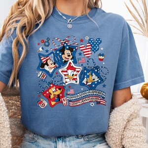 May include: A blue t-shirt with a patriotic design featuring Mickey Mouse, Minnie Mouse, Donald Duck, and Goofy in star-shaped frames. The design includes red, white, and blue elements, balloons, and the text "UNITED STATES OF AMERICA 250TH ANNIVERSARY."