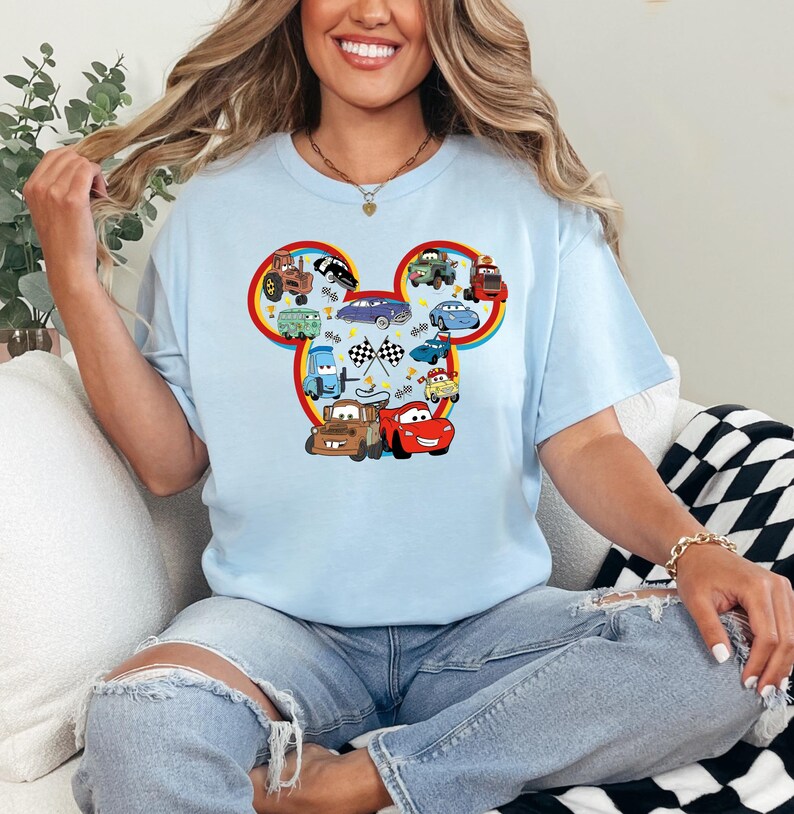 Pode incluir: Uma t-shirt de manga curta azul-clara com um grande gr&aacute;fico em forma de silhueta das orelhas do Mickey Mouse, preenchido com v&aacute;rios carros de desenhos animados do filme Carros, incluindo Rel&acirc;mpago McQueen e Mate, e bandeiras de corrida.