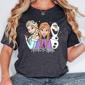 以下が含まれることがあります： アナと雪の女王のエルサ、アナ、オラフのグラフィックプリントが施されたダークチャコールグレーの半袖Tシャツ。キャラクターは白い雪の結晶の列の上に配置されており、生地には小さなカラフルな星が散りばめられています。