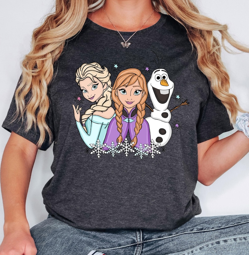 以下が含まれることがあります： アナと雪の女王のエルサ、アナ、オラフのグラフィックプリントが施されたダークチャコールグレーの半袖Tシャツ。キャラクターは白い雪の結晶の列の上に配置されており、生地には小さなカラフルな星が散りばめられています。
