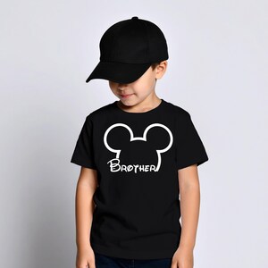 May include: A young child wearing a black baseball cap and a black t-shirt. The t-shirt features a white Mickey Mouse head outline with the word "Brother" printed below it. The child is looking down.