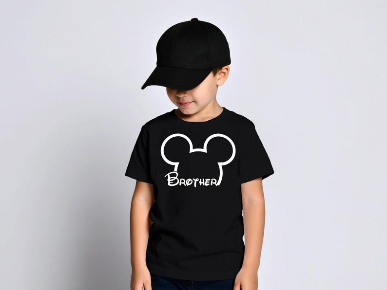 May include: A young child wearing a black baseball cap and a black t-shirt. The t-shirt features a white Mickey Mouse head outline with the word "Brother" printed below it. The child is looking down.