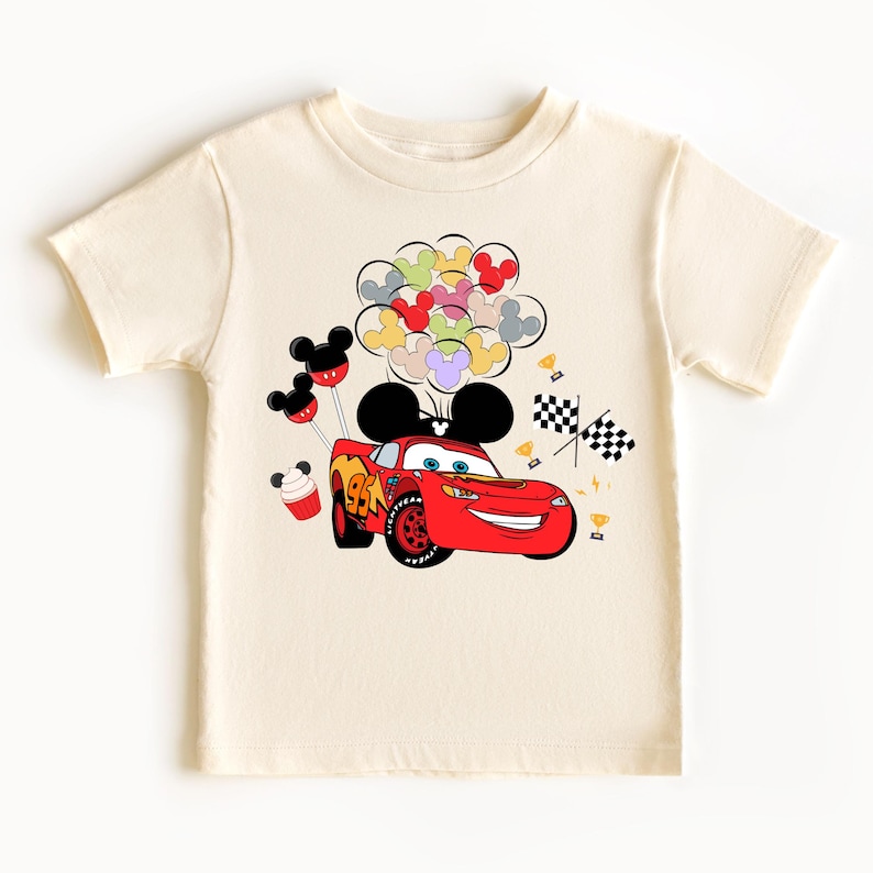 K&ouml;nnte beinhalten: Cremefarbenes kurz&auml;rmeliges T-Shirt mit einem roten Rennwagen mit der Nummer 95, einem l&auml;chelnden Gesicht und Mickey Mouse-Ohren. Das Design umfasst bunte Luftballons, karierte Flaggen und kleine Troph&auml;en.