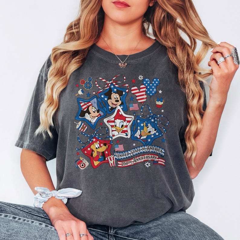 May include: Dark grey t-shirt with a patriotic design featuring Mickey Mouse, Minnie Mouse, Donald Duck, and Goofy in star-shaped frames. The design includes red, white, and blue elements, with text that reads "UNITED STATES OF AMERICA 250TH ANNIVERSARY."