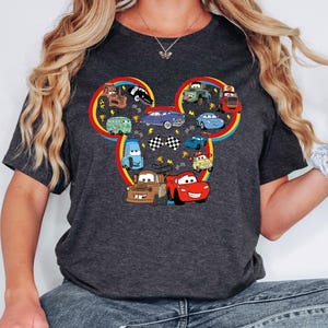 Pode incluir: Uma t-shirt cinzenta escura de manga curta com um grande gr&aacute;fico em forma de silhueta da cabe&ccedil;a do Mickey Mouse, preenchido com v&aacute;rios carros e cami&otilde;es de desenhos animados coloridos, incluindo um carro de corrida vermelho, um reboque castanho e bandeiras axadrezadas.