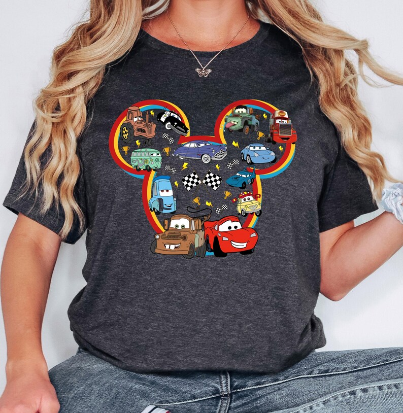 Pode incluir: Uma t-shirt cinzenta escura de manga curta com um grande gr&aacute;fico em forma de silhueta da cabe&ccedil;a do Mickey Mouse, preenchido com v&aacute;rios carros e cami&otilde;es de desenhos animados coloridos, incluindo um carro de corrida vermelho, um reboque castanho e bandeiras axadrezadas.
