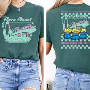 May include: Teal green t-shirt featuring a Pizza Planet design with a rocket, restaurant, and text. The back of the shirt has the text "Interstellar Fast-Food Fueling Station" and three green aliens in blue jumpsuits. The text "Friday Night Kid Night" is also on the back.