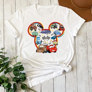 Pode incluir: Uma t-shirt branca de manga curta com um grande gr&aacute;fico em forma de silhueta da cabe&ccedil;a do Mickey Mouse, preenchido com uma colagem de v&aacute;rios carros de desenhos animados coloridos, bandeiras de corrida axadrezadas e pequenos trof&eacute;us dourados.
