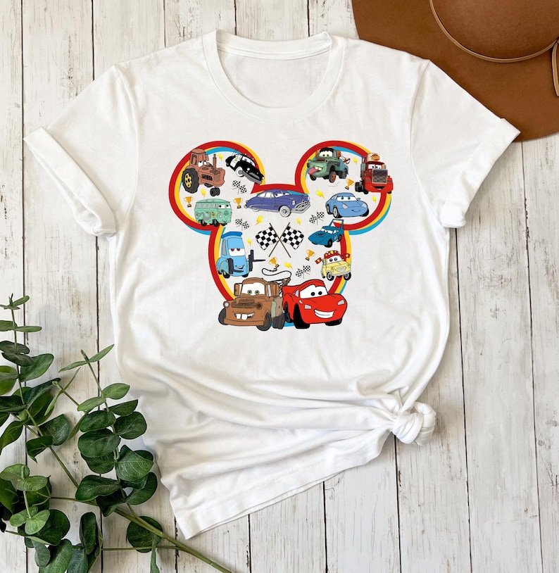 Pode incluir: Uma t-shirt branca de manga curta com um grande gr&aacute;fico em forma de silhueta da cabe&ccedil;a do Mickey Mouse, preenchido com uma colagem de v&aacute;rios carros de desenhos animados coloridos, bandeiras de corrida axadrezadas e pequenos trof&eacute;us dourados.