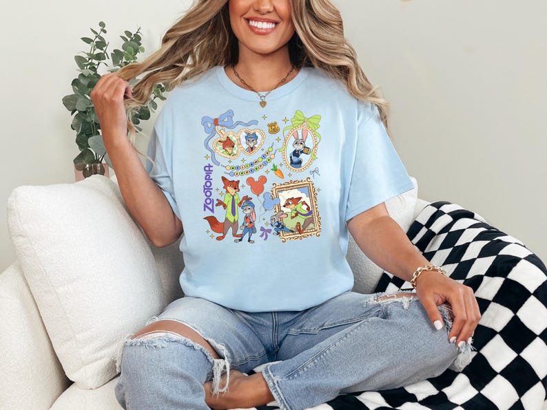 Op de afbeelding: Lichtblauw T-shirt met kleurrijke illustraties van personages uit de film "Zootopia". Het ontwerp bevat ingelijste portretten, personages en het woord "Zootopia" in een verticaal script. Het shirt wordt door een persoon gedragen.