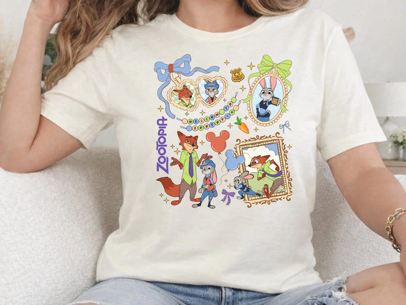 Op de afbeelding: Cr&egrave;mekleurig T-shirt met kleurrijke cartoonillustraties van personages uit de film "Zootopia". Het ontwerp bevat ingelijste portretten, een wortel en het woord "Zootopia" in een verticale lettertype. Het shirt heeft korte mouwen.