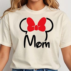 May include: Cream-colored t-shirt featuring a black silhouette of a mouse head with a red bow and the word "Mom" in black script. The shirt has short sleeves and a crew neck.