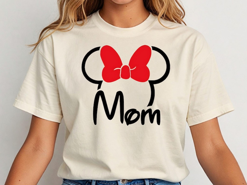 May include: Cream-colored t-shirt featuring a black silhouette of a mouse head with a red bow and the word "Mom" in black script. The shirt has short sleeves and a crew neck.