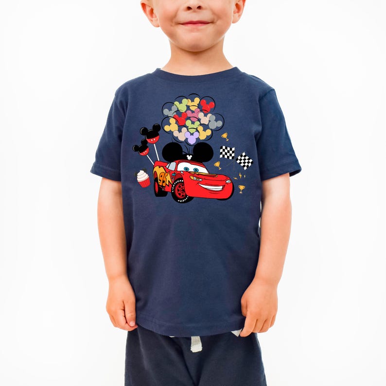 K&ouml;nnte beinhalten: Marineblaues T-Shirt mit einer Cartoon-Grafik eines roten Rennwagens mit einem l&auml;chelnden Gesicht, schwarzen Mickey-Mouse-Ohren und bunten Luftballons. Das Design umfasst karierte Flaggen und einen Cupcake. Das T-Shirt wird von einem Kind getragen.