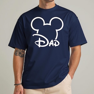 May include: Navy blue t-shirt featuring a white Mickey Mouse head outline with the word "DAD" in a stylized font. The shirt has short sleeves and a crew neck.
