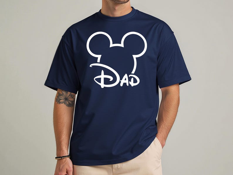 May include: Navy blue t-shirt featuring a white Mickey Mouse head outline with the word "DAD" in a stylized font. The shirt has short sleeves and a crew neck.