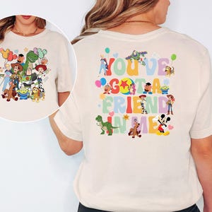 May include: Cream-coloured t-shirt with a colourful graphic of Toy Story characters and the phrase "You've Got a Friend in Me." The design includes Woody, Buzz Lightyear, Rex, and other characters from the animated film.