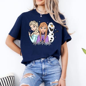 以下が含まれることがあります： 半袖のネイビーブルーのTシャツ。水色のドレスを着た金髪の女性、三つ編みの赤褐色の髪で紫色のケープを羽織った女性、そして白い雪だるまという3人のアニメキャラクターのグラフィックプリントが施されており、小さな星や雪の結晶が描かれています。