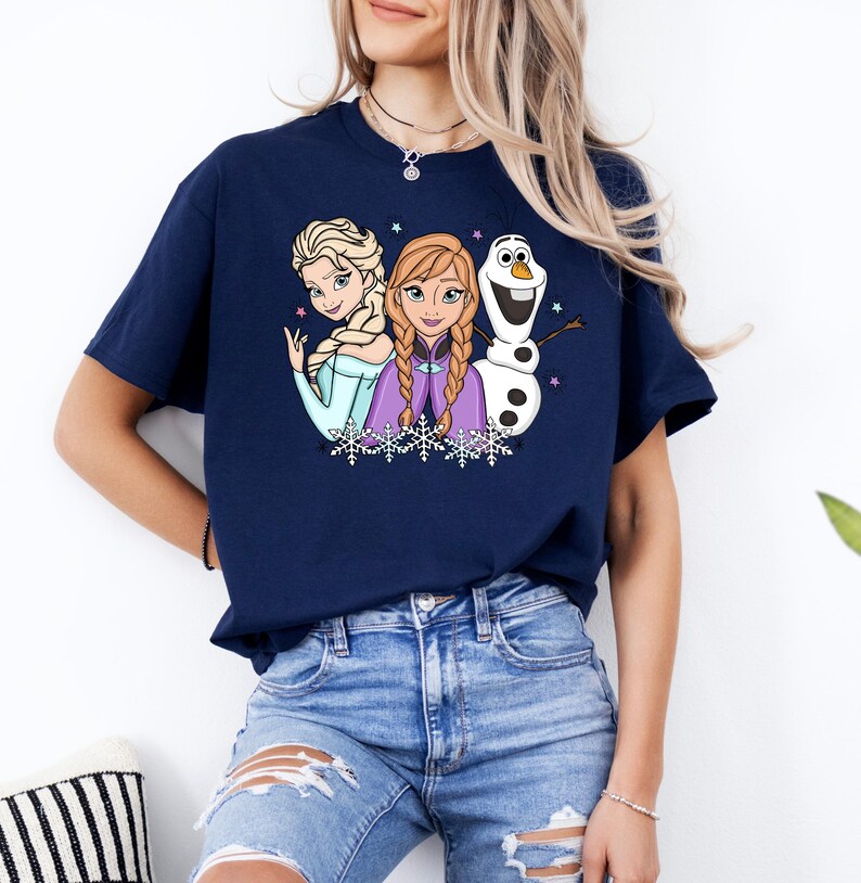 以下が含まれることがあります： 半袖のネイビーブルーのTシャツ。水色のドレスを着た金髪の女性、三つ編みの赤褐色の髪で紫色のケープを羽織った女性、そして白い雪だるまという3人のアニメキャラクターのグラフィックプリントが施されており、小さな星や雪の結晶が描かれています。