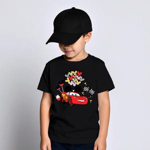 K&ouml;nnte beinhalten: Ein schwarzes T-Shirt mit einem Cartoon-Bild von Lightning McQueen aus dem Film Cars. Das Design umfasst bunte Mickey Mouse-f&ouml;rmige Luftballons, einen Cupcake und karierte Flaggen. Das Kind tr&auml;gt eine schwarze Baseballkappe.