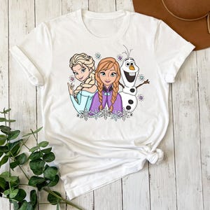 以下が含まれることがあります： 白の半袖Tシャツで、ブロンドと赤褐色の三つ編みヘアの女性キャラクター2人と、白い雪だるまのキャラクターが描かれたカラフルなグラフィックプリントが施されています。デザインには小さな紫と青の星や雪の結晶のアクセントが含まれています。