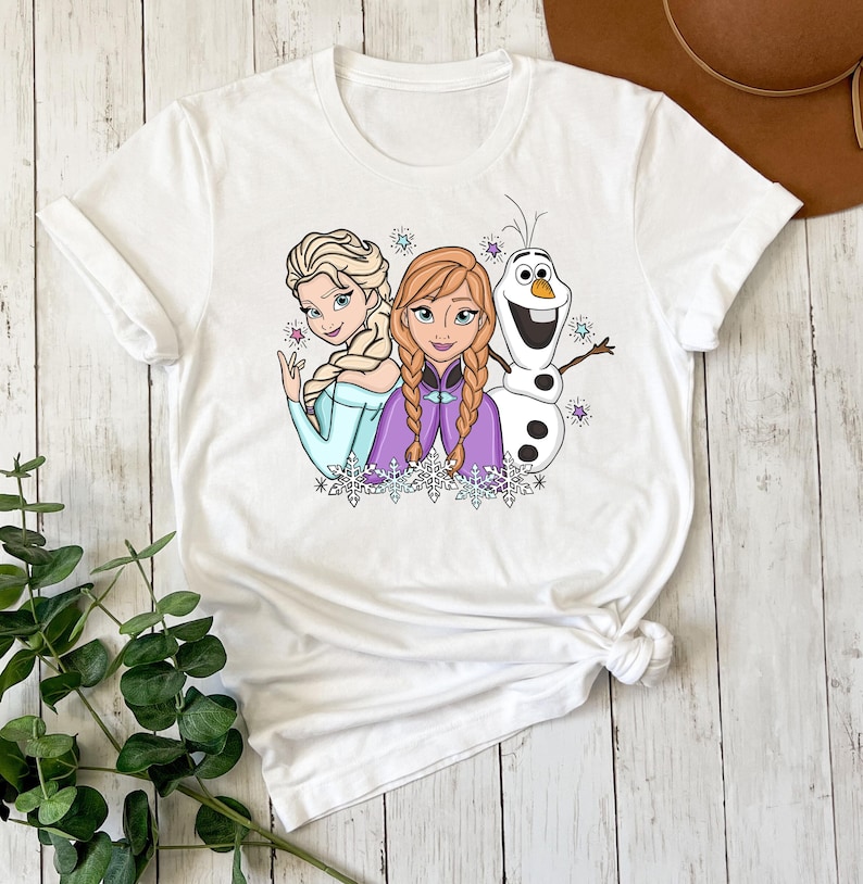 以下が含まれることがあります： 白の半袖Tシャツで、ブロンドと赤褐色の三つ編みヘアの女性キャラクター2人と、白い雪だるまのキャラクターが描かれたカラフルなグラフィックプリントが施されています。デザインには小さな紫と青の星や雪の結晶のアクセントが含まれています。