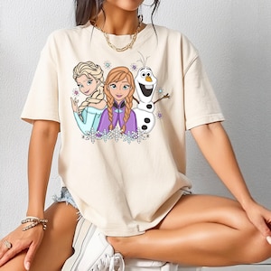 以下が含まれることがあります： 三人のアニメキャラクターが描かれたベージュの半袖Tシャツ。三つ編みのブロンド女性、赤茶色の三つ編みヘアの女性、白い雪だるまがプリントされています。デザインには小さなカラフルな星と雪の結晶があしらわれています。