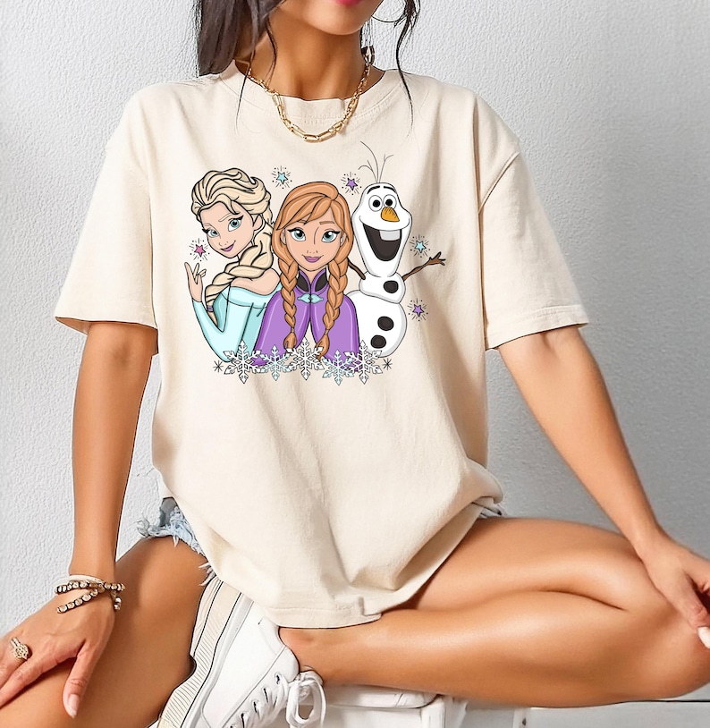 以下が含まれることがあります： 三人のアニメキャラクターが描かれたベージュの半袖Tシャツ。三つ編みのブロンド女性、赤茶色の三つ編みヘアの女性、白い雪だるまがプリントされています。デザインには小さなカラフルな星と雪の結晶があしらわれています。