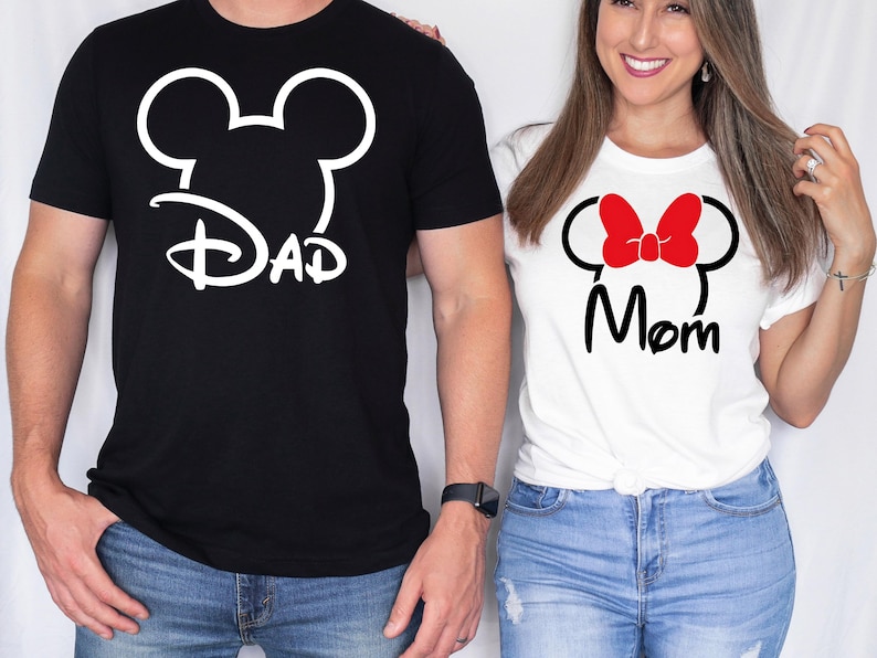 May include: Black t-shirt with a white Mickey Mouse head design and the word "Dad". White t-shirt with a red bow, black Mickey Mouse head design, and the word "Mom". Both shirts are short-sleeved.