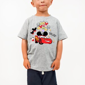K&ouml;nnte beinhalten: Ein graues T-Shirt mit einem roten Cartoon-Rennwagen-Design, komplett mit Mickey Mouse-Ohren und Luftballons. Das Shirt hat auch schwarz-wei&szlig; karierte Flaggen und Mickey Mouse-Themenballons.