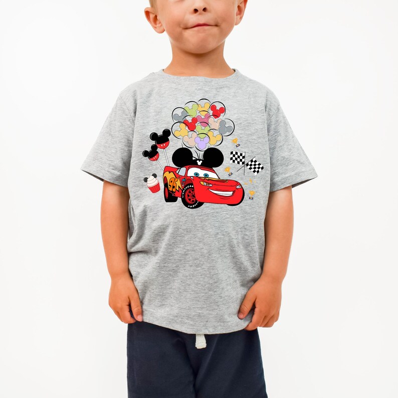K&ouml;nnte beinhalten: Ein graues T-Shirt mit einem roten Cartoon-Rennwagen-Design, komplett mit Mickey Mouse-Ohren und Luftballons. Das Shirt hat auch schwarz-wei&szlig; karierte Flaggen und Mickey Mouse-Themenballons.