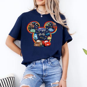 Pode incluir: Uma t-shirt azul-marinho de manga curta com um grande gr&aacute;fico da silhueta da cabe&ccedil;a do Mickey Mouse, preenchido com v&aacute;rios carros de desenhos animados coloridos, bandeiras de corrida axadrezadas e pequenos &iacute;cones de trof&eacute;us dourados, contornado por um arco-&iacute;ris.