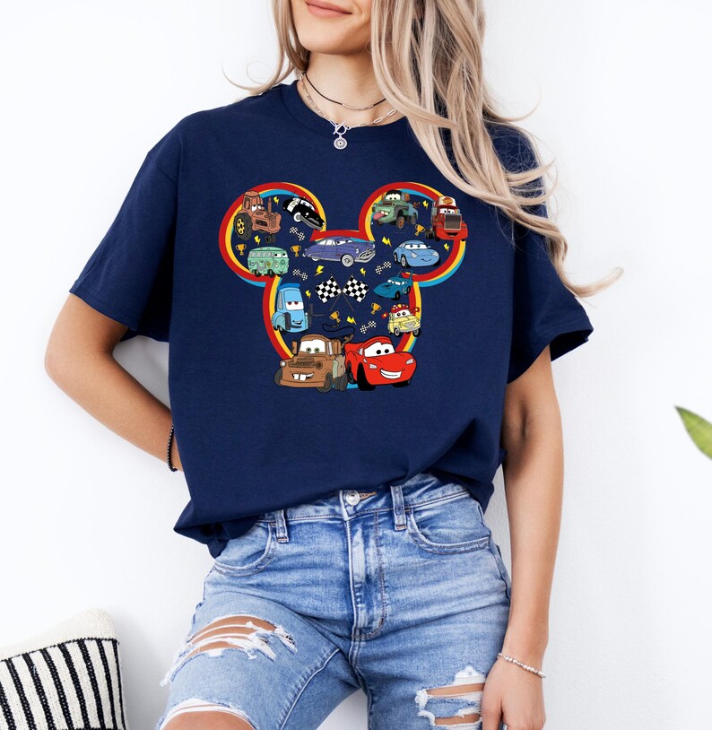 Pode incluir: Uma t-shirt azul-marinho de manga curta com um grande gr&aacute;fico da silhueta da cabe&ccedil;a do Mickey Mouse, preenchido com v&aacute;rios carros de desenhos animados coloridos, bandeiras de corrida axadrezadas e pequenos &iacute;cones de trof&eacute;us dourados, contornado por um arco-&iacute;ris.