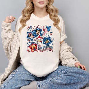 May include: Cream-coloured t-shirt with a patriotic design featuring Mickey Mouse, Minnie Mouse, Donald Duck, and Goofy inside star shapes. The design includes red, white, and blue colours, stars, and the text "United States of America 250th Anniversary."