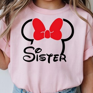May include: A light pink t-shirt featuring a Minnie Mouse design. The design includes a red bow, black mouse ears, and the word "Sister" in a playful font. The shirt is likely for a child.