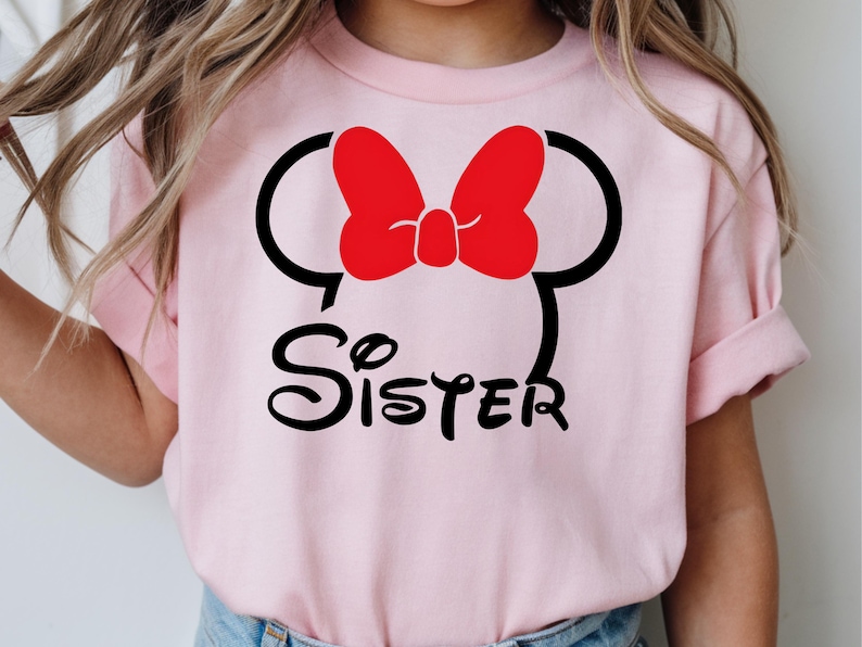 May include: A light pink t-shirt featuring a Minnie Mouse design. The design includes a red bow, black mouse ears, and the word "Sister" in a playful font. The shirt is likely for a child.