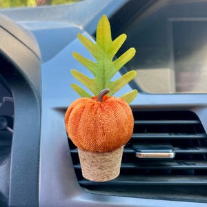 Puede incluir: Un ambientador para coche con forma de calabaza naranja con una hoja verde y amarilla y una base de corcho. La calabaza está hecha de una tela texturizada. El ambientador está diseñado para engancharse a la rejilla de ventilación del coche.
