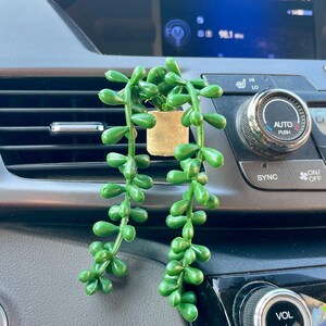 May include: A small, artificial succulent plant in a tan pot, hanging from a car vent. The plant has bright green, teardrop-shaped leaves. The car's dashboard is visible in the background, with radio controls and a volume knob.