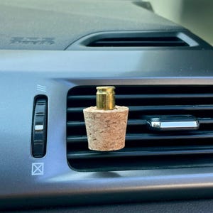 May include: A car air vent diffuser featuring a cork base and a brass bullet casing top. The diffuser is designed to fit into a car's air vent. The cork base is cylindrical and has a natural, textured appearance.