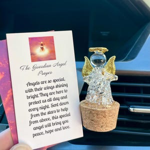 May include: A clear glass angel figurine with gold glitter wings and halo, atop a cork base. A card with the text "The Guardian Angel Prayer" is held nearby. The angel is a decorative item.