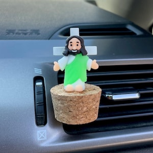 Pode incluir: Um clipe de ventilação de carro com uma pequena figura de Jesus com uma túnica verde, mangas brancas e uma cruz. A figura é montada em uma base de cortiça. O fundo mostra o painel de um carro.