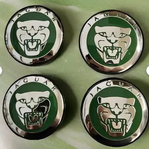 Op de afbeelding: Vier ronde Jaguar-auto-emblemen. Elk embleem heeft een zilveren jaguar-kop op een groene achtergrond, omgeven door een zilveren ring met het woord "JAGUAR" in zwarte letters. De emblemen hebben een zwarte basis.