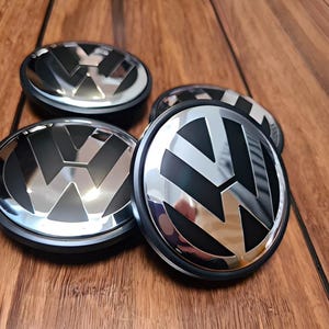May include: Four chrome and black Volkswagen hubcaps with the VW logo. The hubcaps are circular with a shiny silver finish on the logo and a black background. They are arranged on a wooden surface.