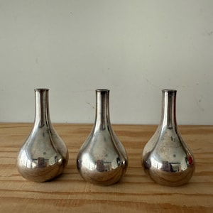 Puede incluir: Tres jarrones plateados en forma de lágrima con cuellos estrechos están dispuestos sobre una superficie de madera. Los jarrones tienen un acabado metálico reflectante, y la luz se refleja en sus superficies curvas. El fondo es una pared lisa de color blanquecino.