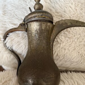 Puede incluir: Cafetera antigua de metal con un diseño floral decorativo y un asa curva. La cafetera tiene un pico largo y una tapa con un pequeño pomo. El metal tiene un aspecto deslustrado y envejecido.