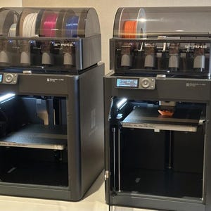 Könnte beinhalten: Zwei schwarze 3D-Drucker mit transparenten Filament-Lagereinheiten. Die Drucker haben offene Türen und beleuchtete Druckbetten. Ein Drucker druckt ein orangefarbenes Objekt. Die Drucker stehen auf einer hellen Oberfläche.